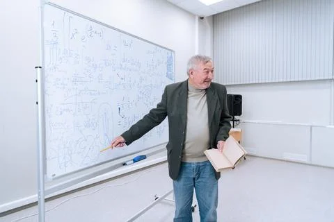 A gray-haired famous professor in a jacket and jeans shows his way of solving Stock Photos