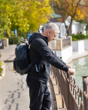 A gray-haired man with a backpack stands on the city embankment and looks at the Stock Photos