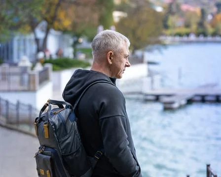 A gray-haired man with a backpack stands on the city embankment and looks at the Stock Photos
