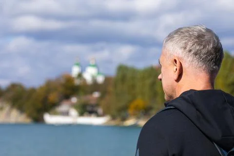 A gray-haired man with a backpack stands on the city embankment and looks at the Stock Photos