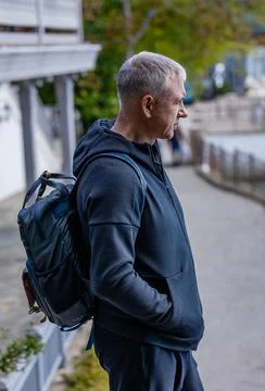 A gray-haired man with a backpack stands on the city embankment and looks at the Stock Photos