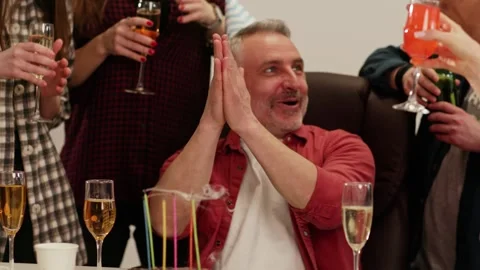 A gray-haired man in a festive cap sits in a circle of friends and blows out Stock Footage 243366784