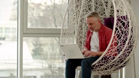 A gray-haired man sits in a hanging chair and types on a laptop. A man works Stock Footage 243367493