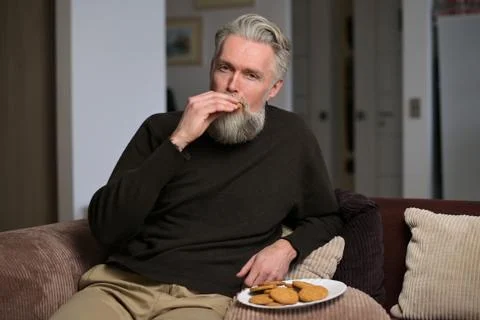 Gray-haired man sitting on the couch at home and eating cookies Stock Photos