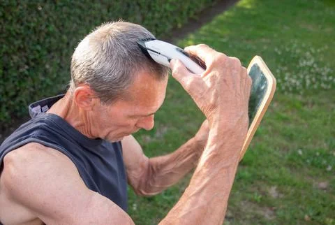 A gray-haired middle-aged man shaves his hair with a clipper in a garden Stock Photos