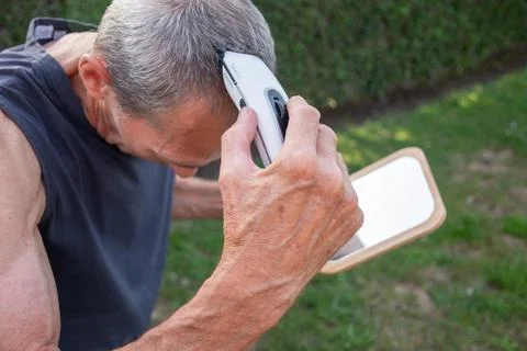 A gray-haired middle-aged man shaves his hair with a clipper in a garden Stock Photos