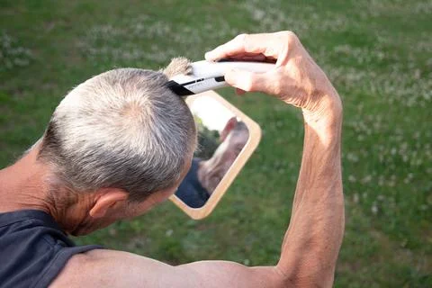 A gray-haired middle-aged man shaves his hair with a clipper in a garden Stock Photos