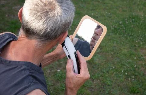 A gray-haired middle-aged man shaves his hair with a clipper in a garden Stock Photos