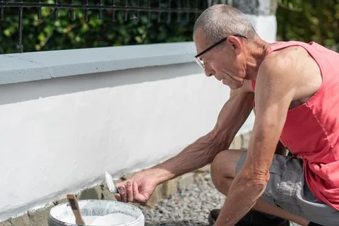 A gray-haired middle-aged man picks up white paint from a bucket with a brush to Stock Photos
