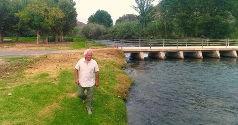 Gray-haired old man walking relaxed on river bank at sunset Stock Footage 124384310