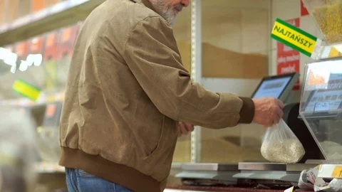 Gray-haired retiree with a beard weighs cereals on scales in a supermarket Stock Footage 99934965