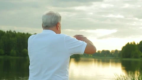 A gray-haired senior man in a white shirt is training on the river bank. Stock Footage 169065824