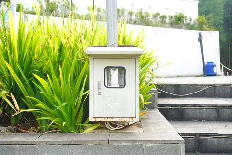 Gray outdoor electrical utility box mounted on concrete sidewalk near green.. 스톡 사진