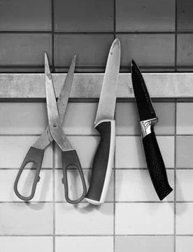 Grayscale shot of scissors and two kitchen knives hanging on the wall Stock Photos