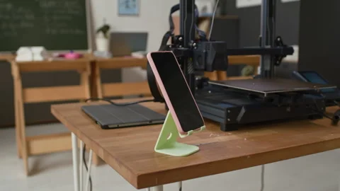 Green 3D Printed Stand Holding Pink Smartphone on Wooden Table in Classroom Stock Footage 335699676