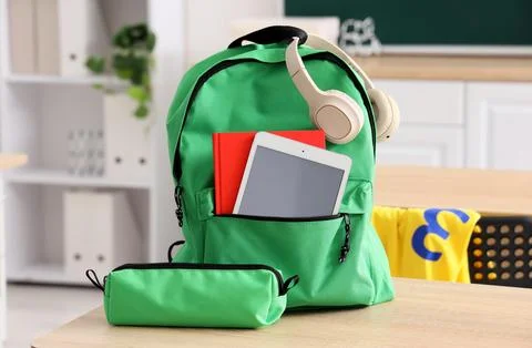 Green backpack with digital tablet, red book, and headphones on a wooden desk Stock Photos