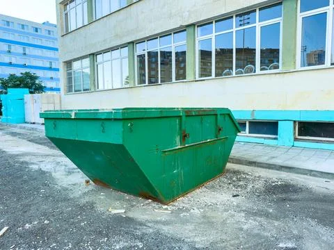 A green dumpster sits next to a building on a city street. The pavement is vi Stock Photos