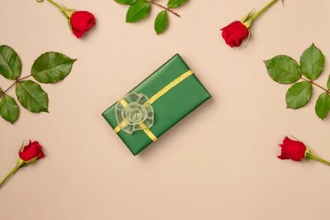 Green gift box in a frame of red roses on beige background from above. Flat.. Stock Photos