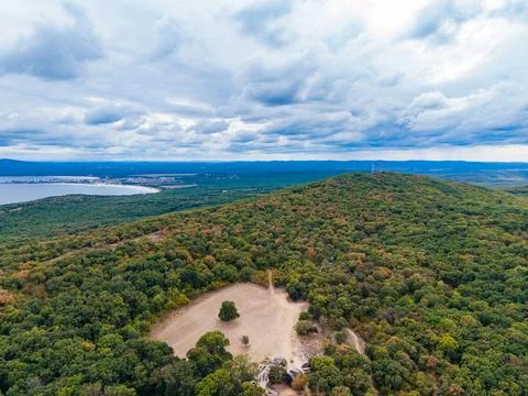 Green hill with trees under cloudy sky near a body of water and distant cit.. Stock Photos