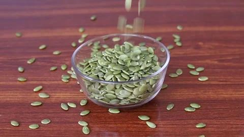 Green hulled pumpkin seeds falling into a clear glass bowl Stock Footage 346444534