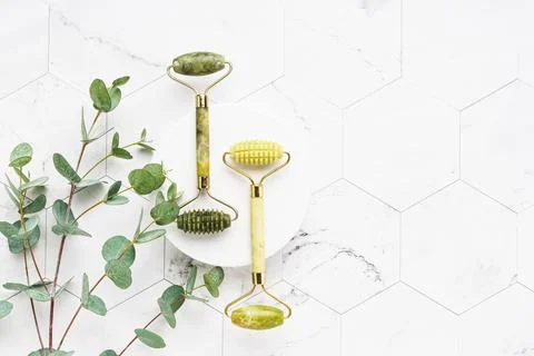Green jade face rollers and eucalyptus branches on a white marble background. Stock Photos