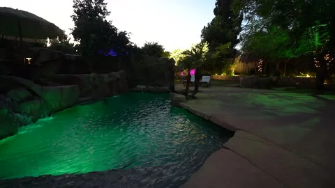 Green lit rock lagoon pool with waterfall in a backyard at dusk Stock Footage 345536550