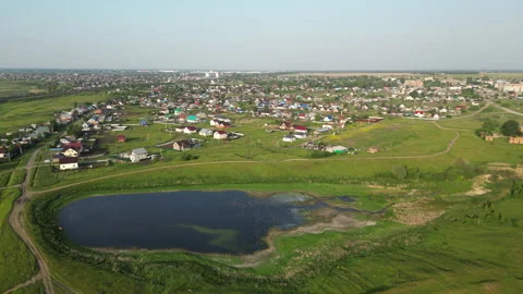 Green meadows surround a tranquil pond near a residential area with colorful Vídeos de archivo 318311708