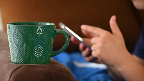 Green mug resting on couch armrest while person scrolls smartphone Stock Footage 339660963