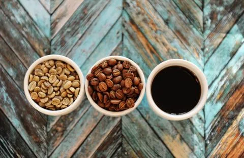 Green raw beans, roasted coffee beans and black coffee in three white cups on Stock Photos