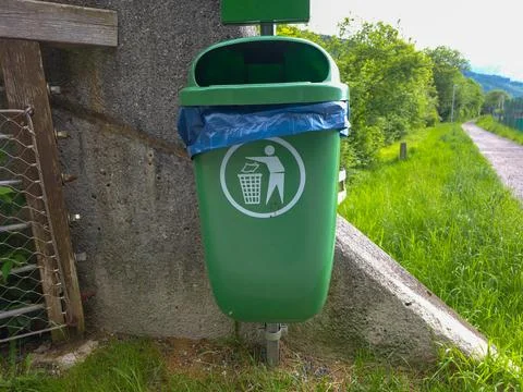 A green recycling bin stands by a path with a concrete wall and a recycling.. Stock Photos