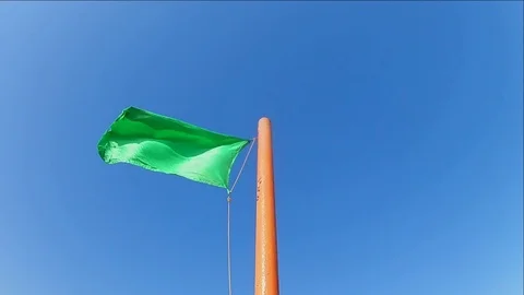 Green safe flag waving on a flagpole against a blue clear sky. Slow motion Timel Stock Footage 114645561