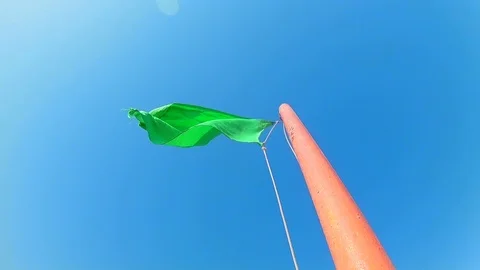 Green safe flag waving on a flagpole against a blue clear sky. Slow motion Ti Stock Footage 115070095