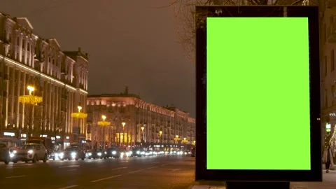 A green screen billboard located on the sidewalk on which people walk, next to a Stock Footage 148311714