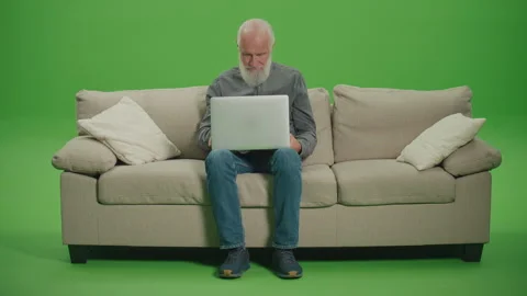 Green Screen. A Smiling Old Man Sitting on a Sofa and Works on a Laptop. Video stock 263396074