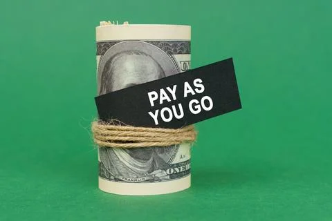 On a green surface, rolled dollars with a black sign that says - PAY AS YOU G Stock Photos