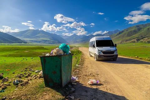 Green trash bin overflowing with garbage on rural dirt road Stock Photos