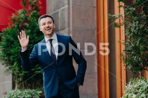 Greeting concept. Cheerful successful young businessman in formal clothes wav Stock Photos