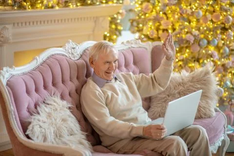 Grey-haired elderly man sitting on the sofa and having a video chat Stock Photos