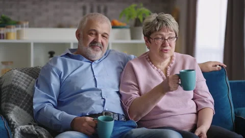 Grey haired man is embracing his aged wife in living room, spouses are sitting Stock Footage 153519114
