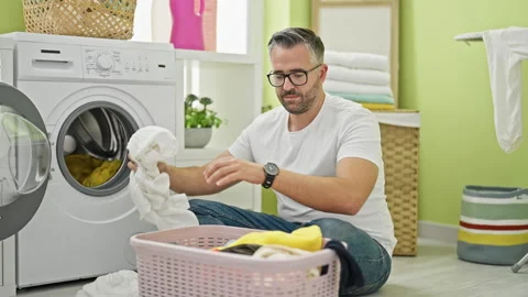 Grey-haired man separating white and color clothes at laundry room Video stock 246992667