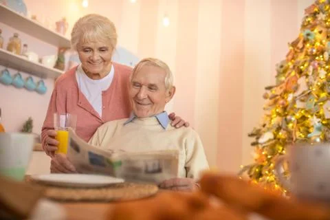 Grey-haired man sitting at the table while his wife giving him orange juice Stock Photos