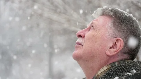 A grey-haired man stands under snowflakes and looks up in a park in winter Stock Footage 85627345