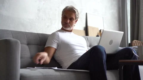 Grey-haired Mature handsome man sitting on sofa and typing on laptop at home Stock Footage 141184013