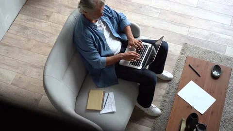 Grey-haired Mature handsome man sitting on sofa and typing on laptop at home Stock Footage 141184142