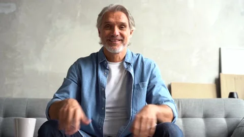 Grey-haired Mature man vlogger look at camera recording blog sit on sofa at home Stock Footage 141192875