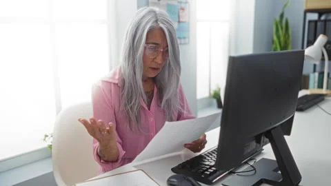 Grey-haired woman working at computer in office, reading documents with foc.. Stock Footage 281131453