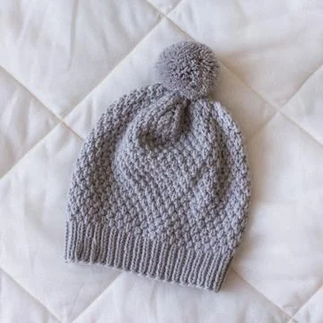 Grey knitted hat with a small pompom on a white blanket Stock Photos