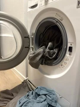 Grey laundry hangs out of an open front loading washing machine with clothes  Foto stock