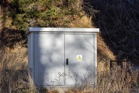A grey metal electrical utility box sits in a field near a forest, displaying a Stock Photos