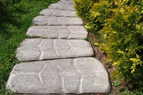 Grey path of embossed patterned shaped concrete slabs lying among green shrubs. Stock Photos